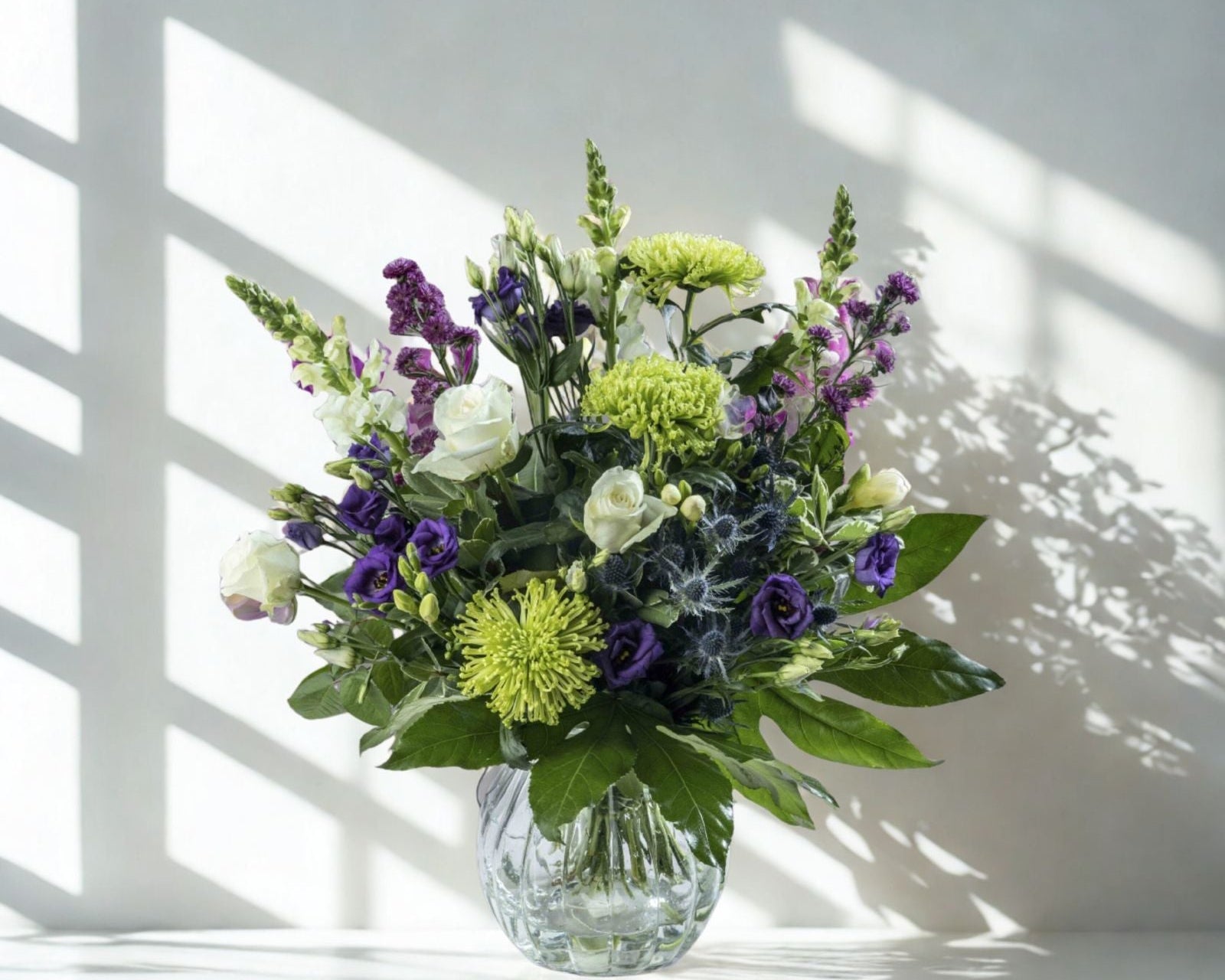 a green, purple and white flower bouquet that is perfect for any occasion especially birthdays, it's on a white reflecting surface with diffused soft light in a glass vase