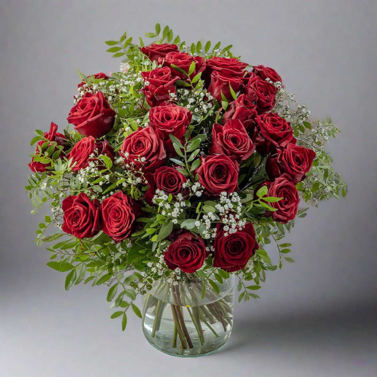 A bouquet of two dozen red roses arranged in a clear vase, with green leaves and white flower accents.