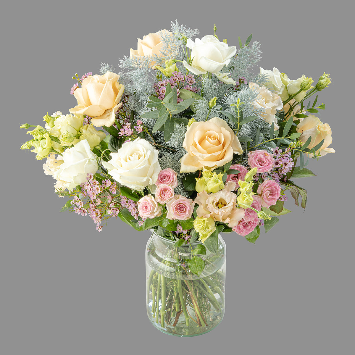 A pastel-colored bouquet of flowers including peach melba and pristine white roses, pink wax flowers, and other greenery in a clear vase.