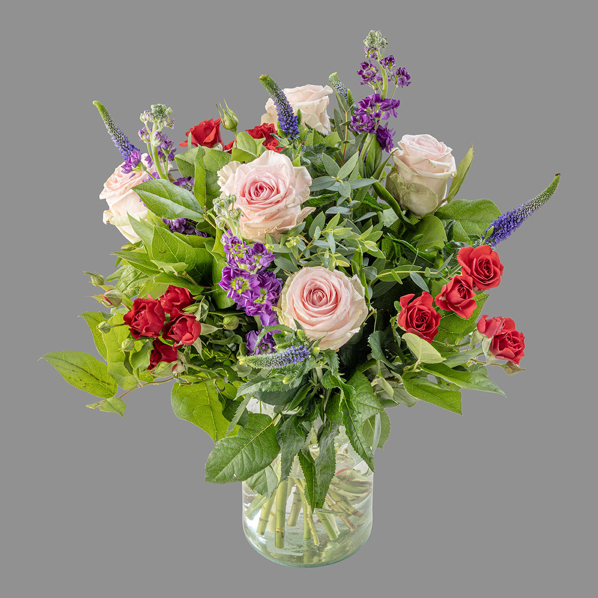A bouquet of flowers featuring pink and red roses, purple flowers, and greenery in a clear vase.