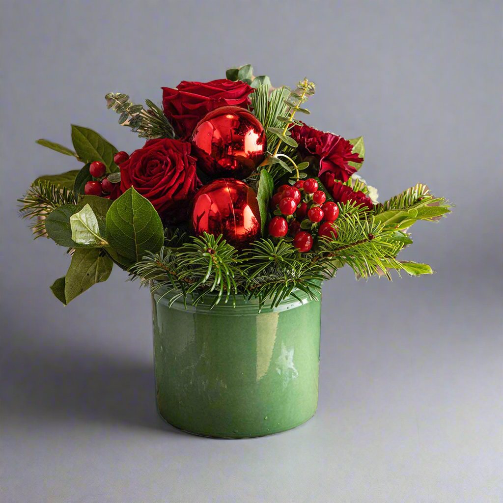 A festive Christmas-themed tabletop arrangement featuring a green container, adorned with a variety of red and green plants including carnations, roses, eucalyptus, and pine needles.