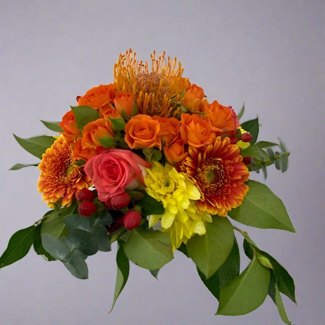 A vibrant orange floral arrangement featuring a variety of flowers including roses and gerberas, wrapped in green foliage.