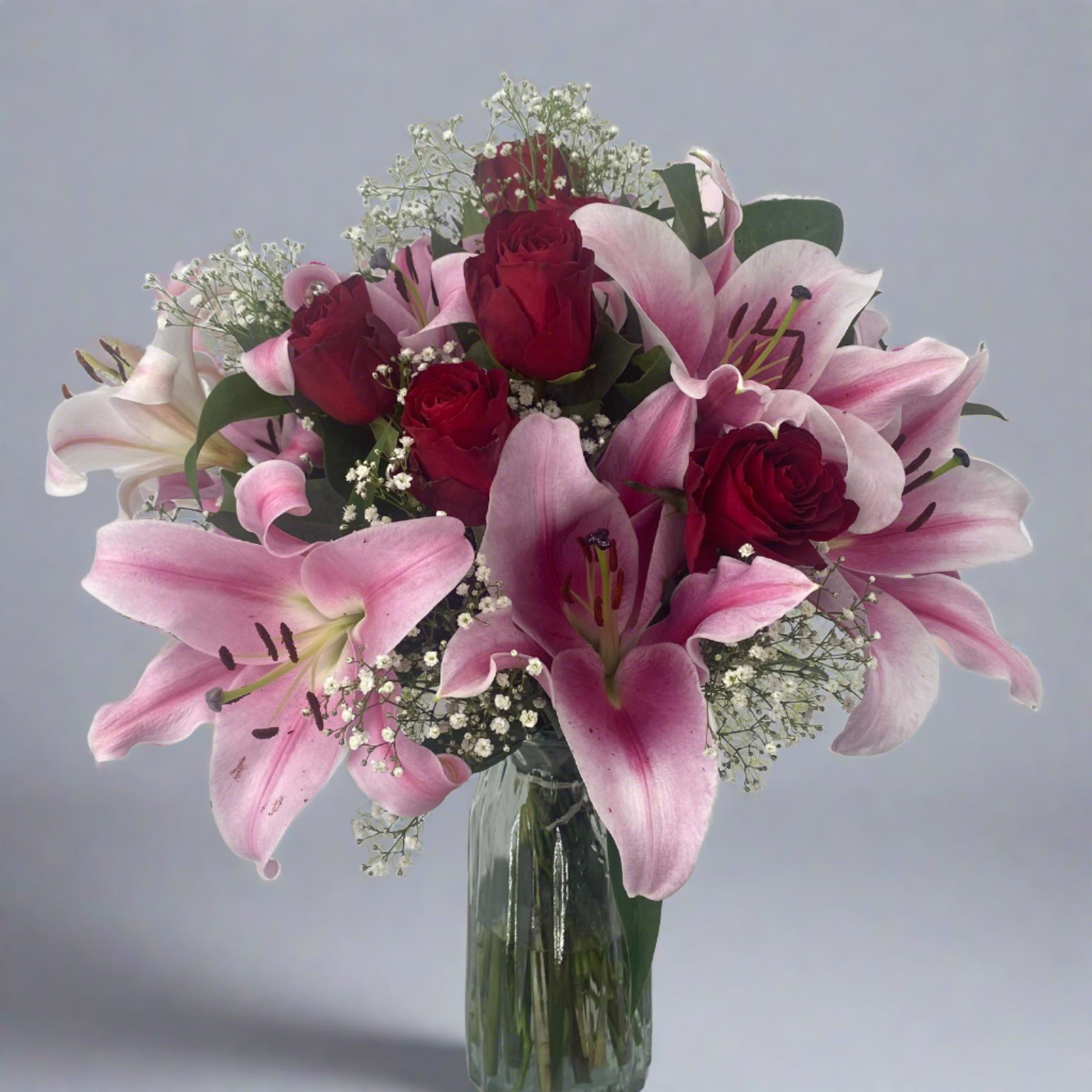 A bouquet of red roses and pink lilies arranged in a clear vase, complemented by green foliage and small white flowers.