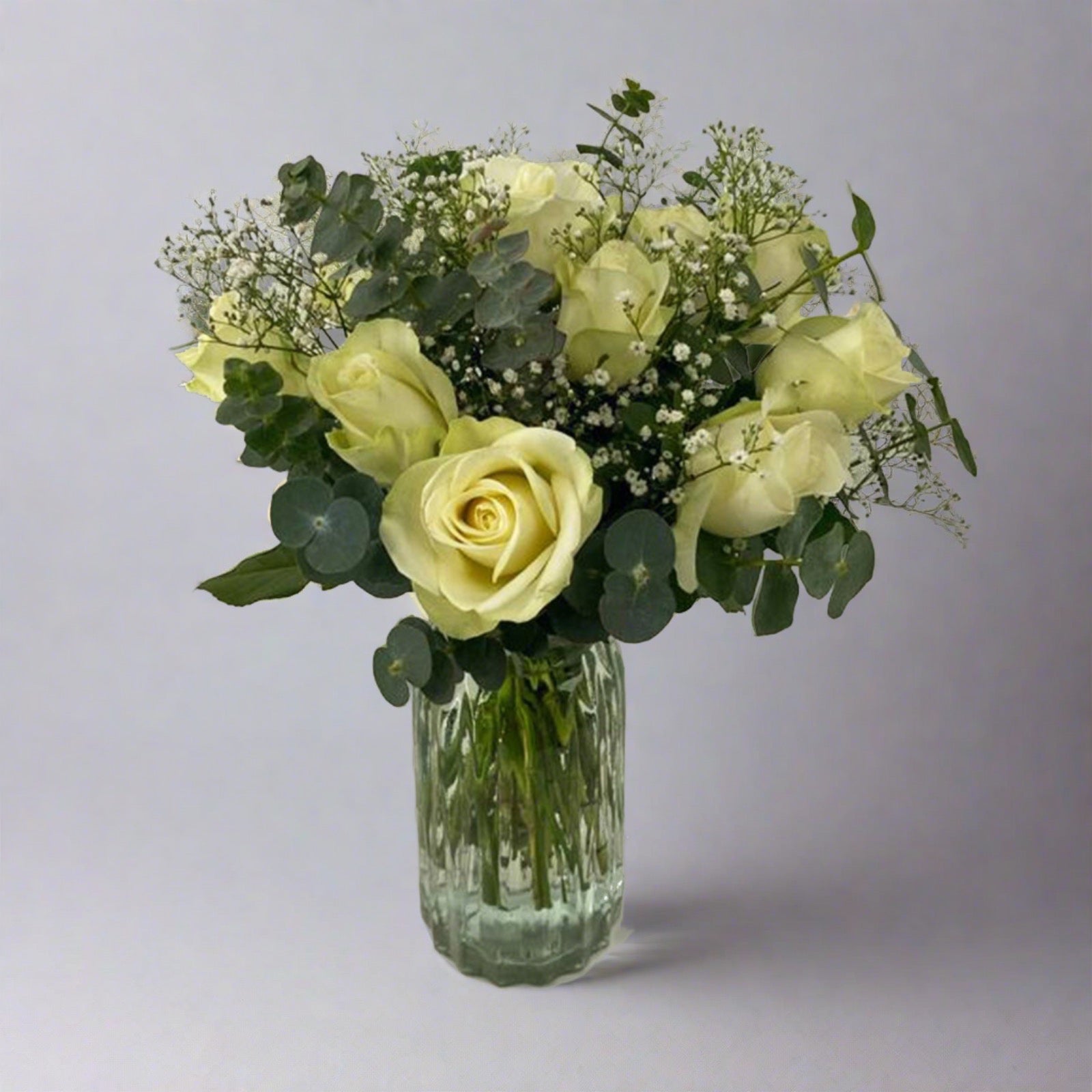 A clear glass vase containing a dozen white roses with green leaves and a few small green flowers, presenting an elegant and understated floral arrangement.