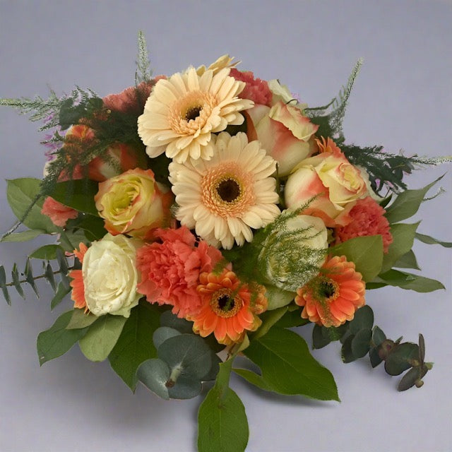 A floral table topper arrangement featuring a variety of flowers including roses and carnations in shades of peach, orange, and cream, with green foliage.
