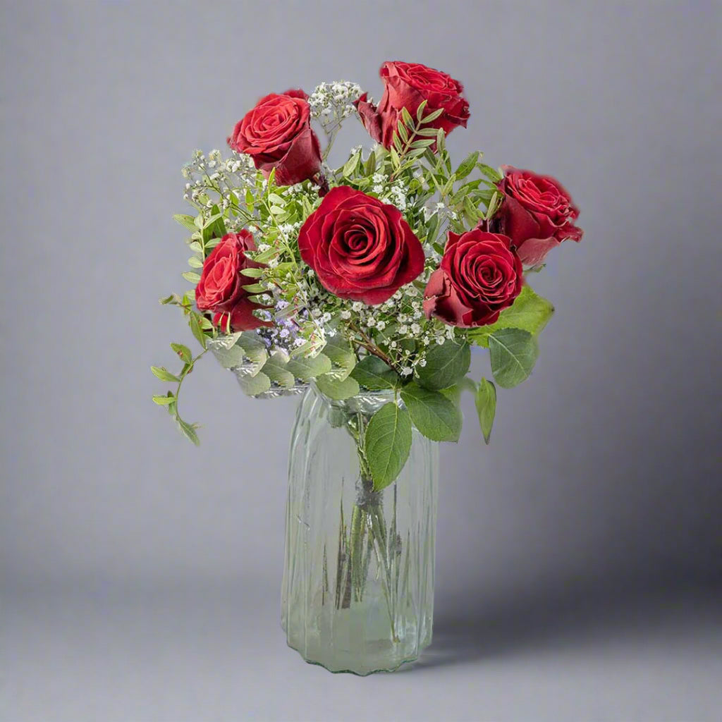 A bouquet of red roses with green leaves, arranged in a clear glass vase.