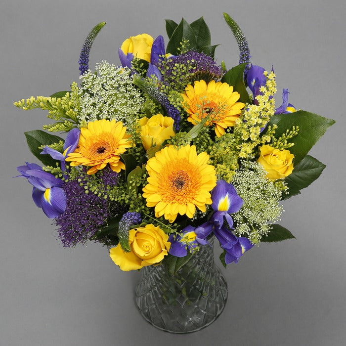 A vibrant Easter flower bouquet containing yellow and purple flowers, along with greenery and filler in a clear vase.
