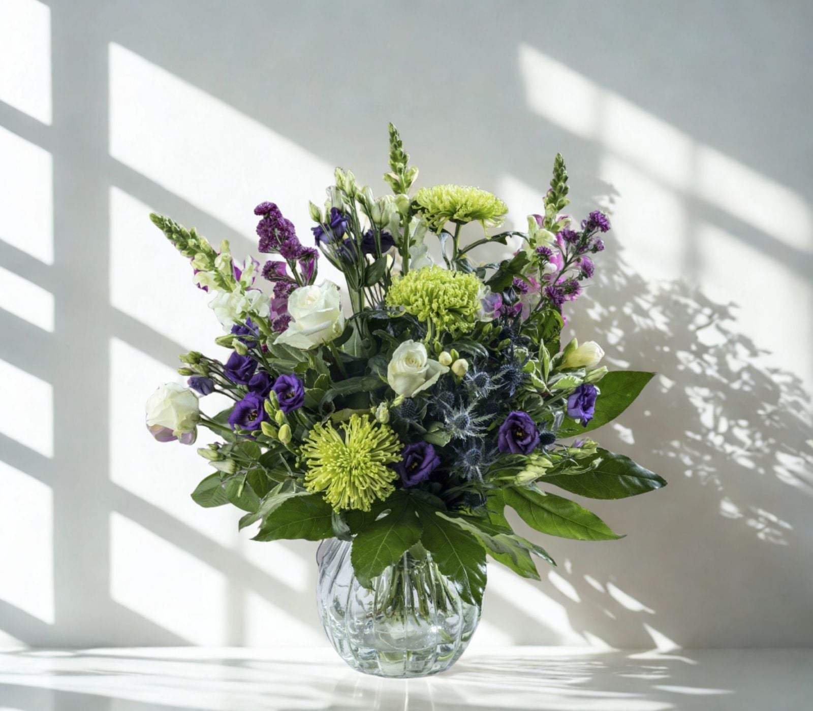 a green, purple and white flower bouquet that is perfect for any occasion especially birthdays, it's on a white reflecting surface with diffused soft light in a glass vase