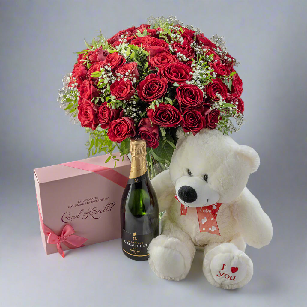 A gift set including a bouquet of red roses, a white teddy bear wearing a red bow, a bottle of champagne, and a box of chocolates, all arranged on a surface.