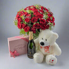 A gift set including a bouquet of red roses, a white teddy bear wearing a red bow, a bottle of champagne, and a box of chocolates, all arranged on a surface.