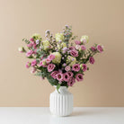 A mixed floral bouquet featuring pink roses, pale yellow lisianthus, and green foliage arranged in a clear ribbed glass vase against a plain white background.
