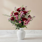 A vibrant floral bouquet featuring deep red and light pink roses, pink lisianthus, and green eucalyptus stems, arranged in a clear ribbed glass vase against a plain white background.