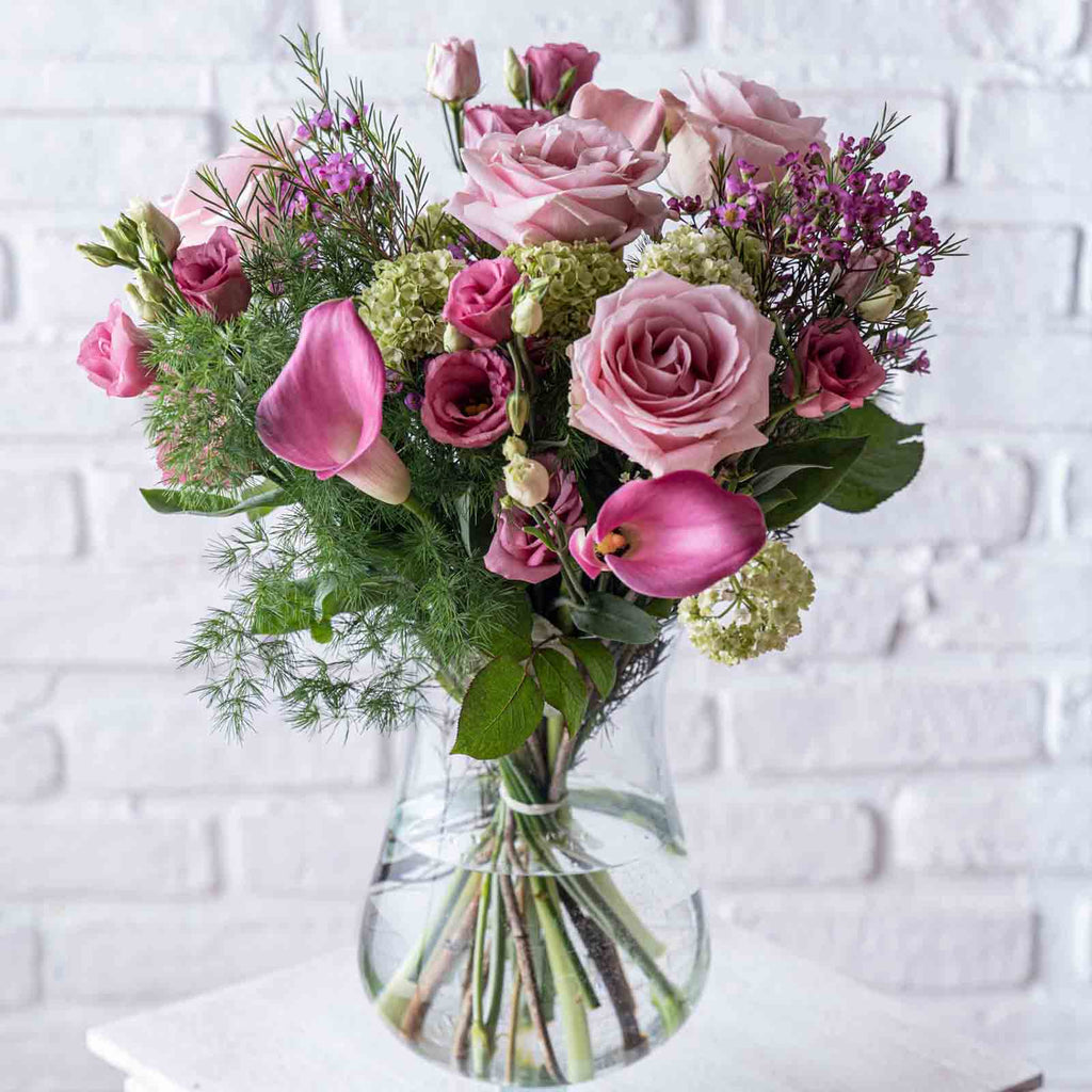 Blush flower bouquet with pink roses in a glass vase against a white brick wall background